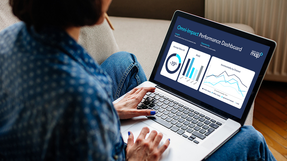 A person sits indoors using a laptop displaying the OmniImpact Performance Dashboard. The screen shows various data visualizations, including bar graphs, pie charts, and line graphs.