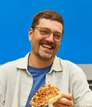 White man with glasses smiling and holding a slice of pizza against a blue background.