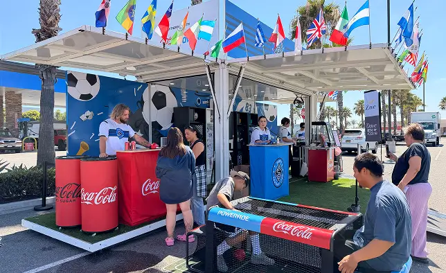 An outdoor promotional booth features Coca-Cola and Powerade counters decorated with soccer graphics and international flags. Several people interact with staff members, receiving drinks and engaging with the display in a sunny parking lot setting. The booth includes colorful national flags overhead, branded coolers, and a small turf area that enhances the sporty atmosphere. Palm trees and parked vehicles in the background suggest a casual, public event location.