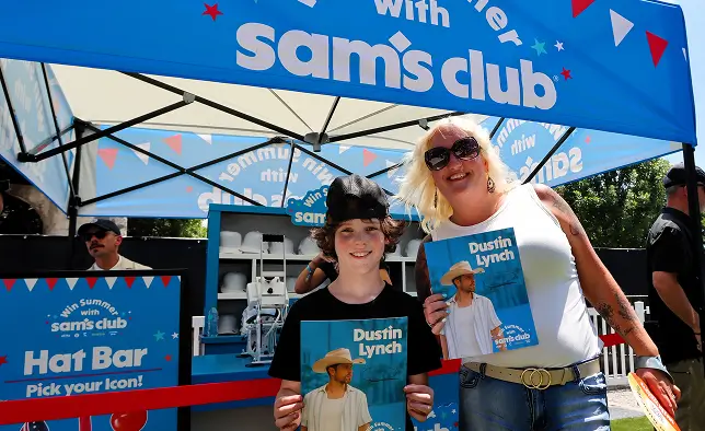 A smiling woman and child pose in front of a bright blue Sam's Club event tent. They each hold a Dustin Lynch poster while standing behind a red rope barrier. The tent features text promoting a summer hat bar and icon selection, with colorful bunting graphics. Other attendees and event staff are visible in the background under the canopy.