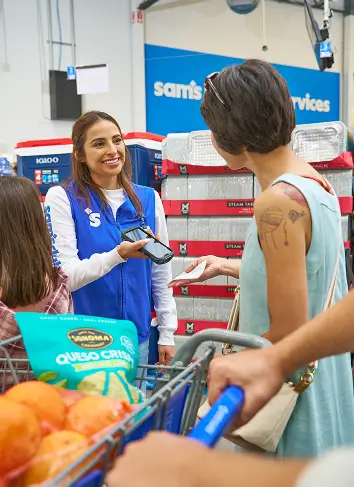 A smiling store associate in a blue vest helps a customer at the checkout area of a warehouse-style retail store. The associate holds a handheld scanner while speaking with a woman standing beside a shopping cart. The cart is filled with groceries, including oranges and a large green product bag, and other shoppers are partially visible in the foreground. In the background, stacked merchandise and a service sign contribute to the busy retail environment.