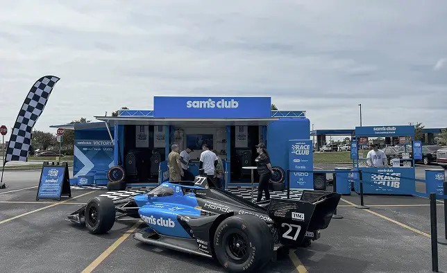 A blue and black Indy-style race car is parked in front of a mobile Sam's Club promotional trailer. Several people stand near the car and trailer, engaging with the display in an outdoor parking lot. The trailer and surrounding signage feature bold blue graphics, checkered racing patterns, and the Sam's Club name. The scene conveys a motorsport-themed marketing activation under a cloudy daytime sky.