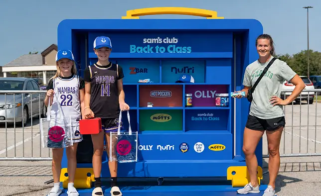 Three people stand in front of a large blue Sam's Club Back to Class display in a parking lot. Two children hold clear shopping bags and a red lunchbox, while an adult stands beside them with a snack item. The display features product panels for ZBAR, Nutri, Hot Pockets, OLLY, and Mott's with bright, colorful graphics. Metal barriers, parked cars, and a building are visible in the background under a clear sky.