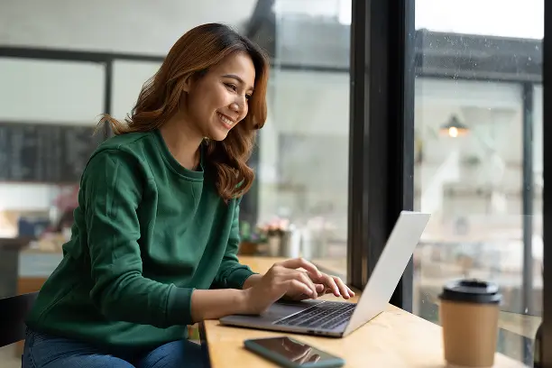 A young woman sits at a wooden table using a laptop near large windows. She smiles as she types, suggesting she is working or studying in a relaxed cafe setting. A takeaway coffee cup and smartphone rest on the table beside her. Soft natural light and warm tones create a comfortable, productive atmosphere.